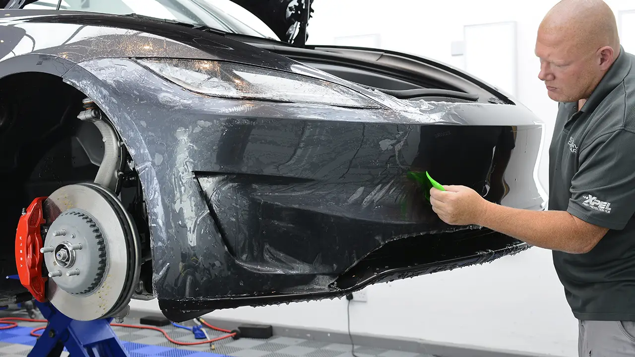 Technician squeegeeing XPEL paint protection film onto the front bumper of a black sports car with the wheel removed